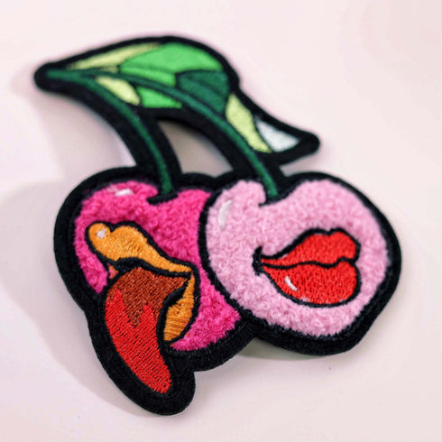 Patch featuring two cherries with expressive lips, one with a tongue sticking out, set against a white background. The design is playful and vibrant, combining bold colors with a fun, cheeky style.